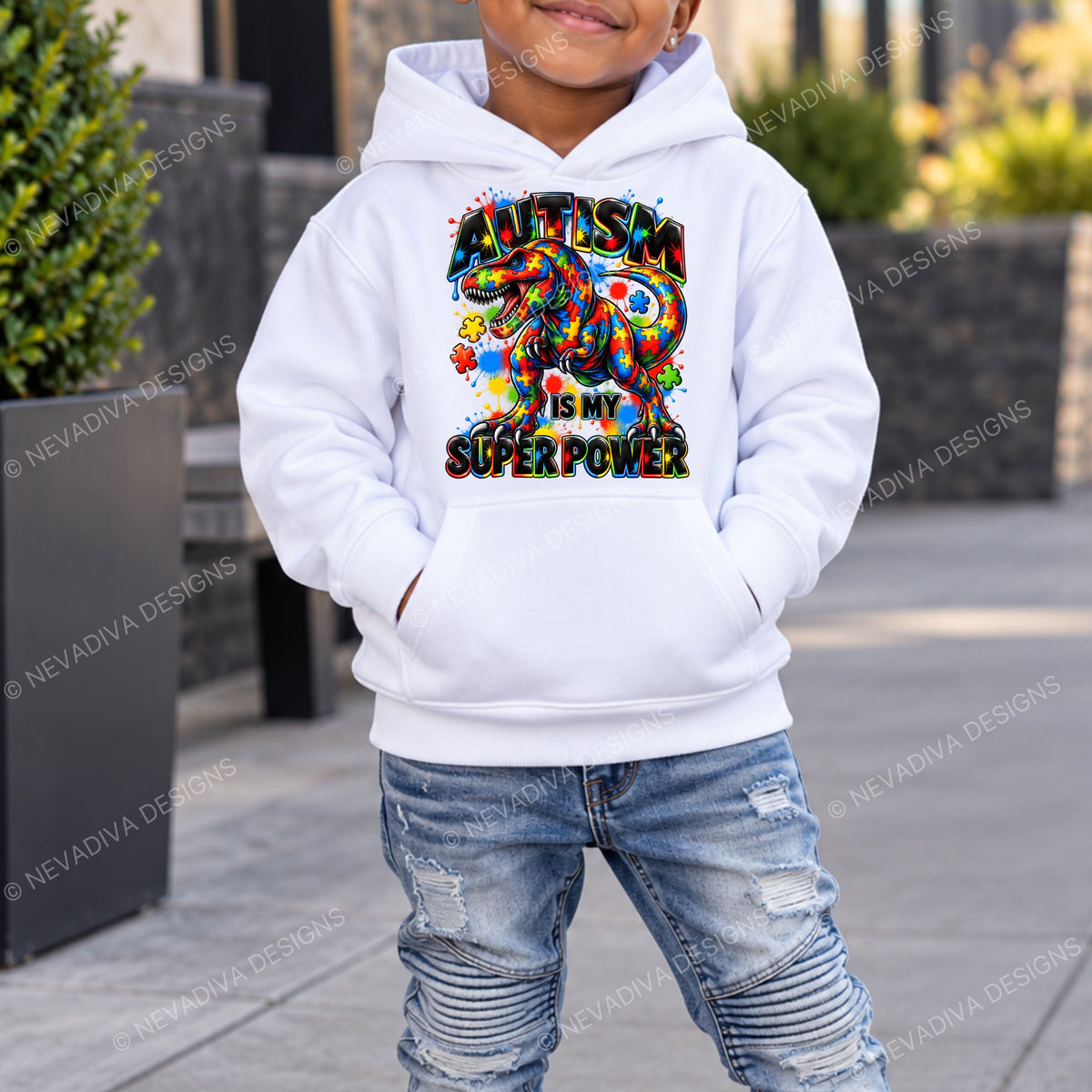 A child wearing a white pullover hoodie with a colorful graphic that reads 'Autism is my Super Power' and distressed blue jeans, standing outdoors on a paved walkway.
