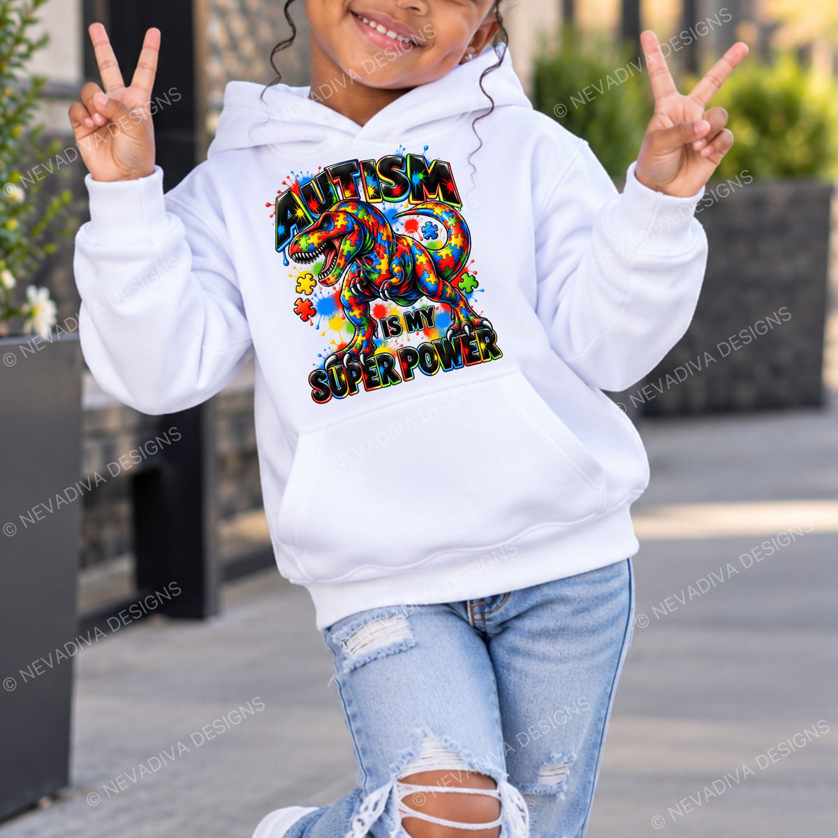 A child wearing a white pullover hoodie with a colorful autism awareness graphic on the front that reads 'Autism is my Super Power,' paired with light blue ripped jeans, posing outdoors with peace signs.