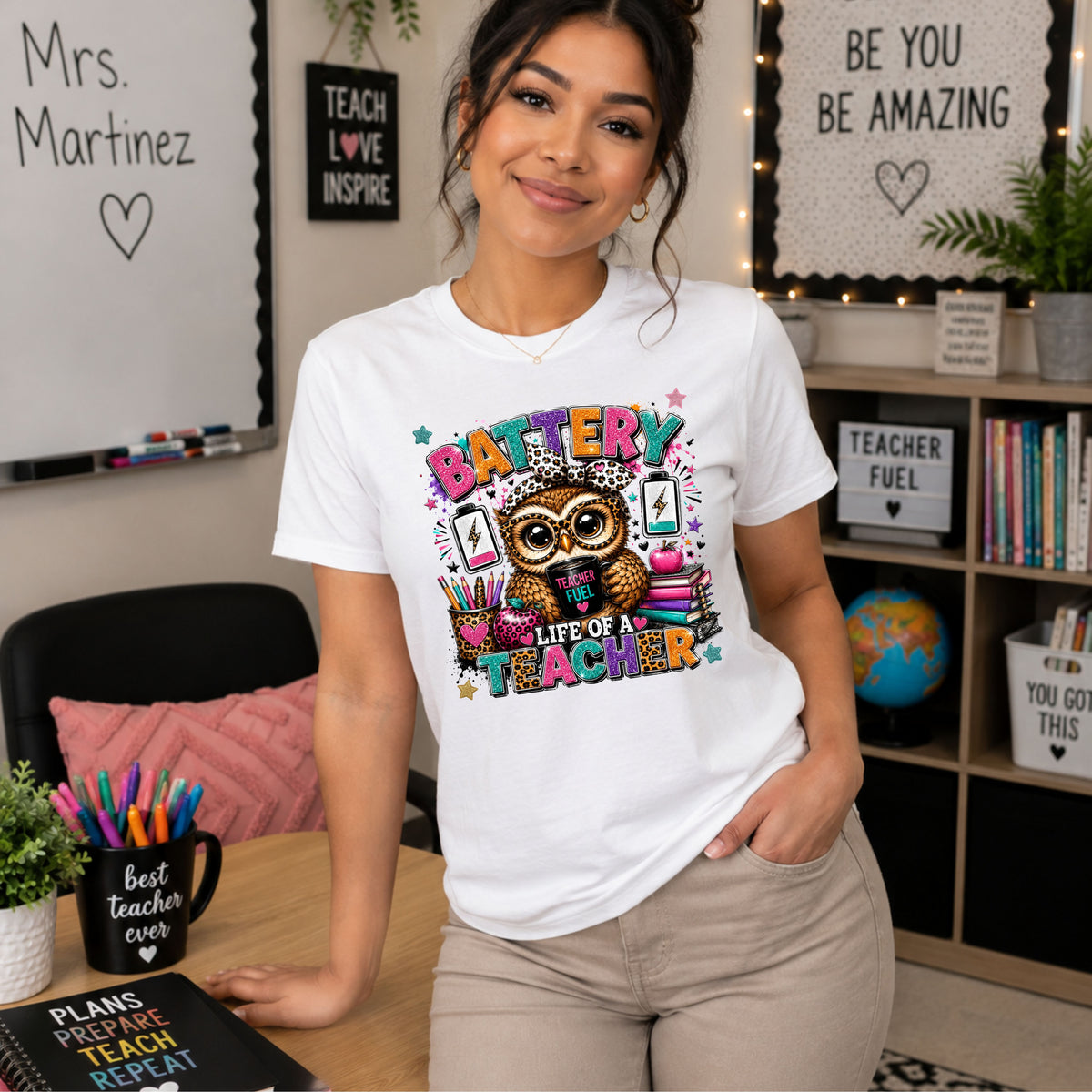 A woman wearing a white graphic t-shirt with a colorful owl and 'BATTERY TEACHER' design, standing in a decorated teacher's room.