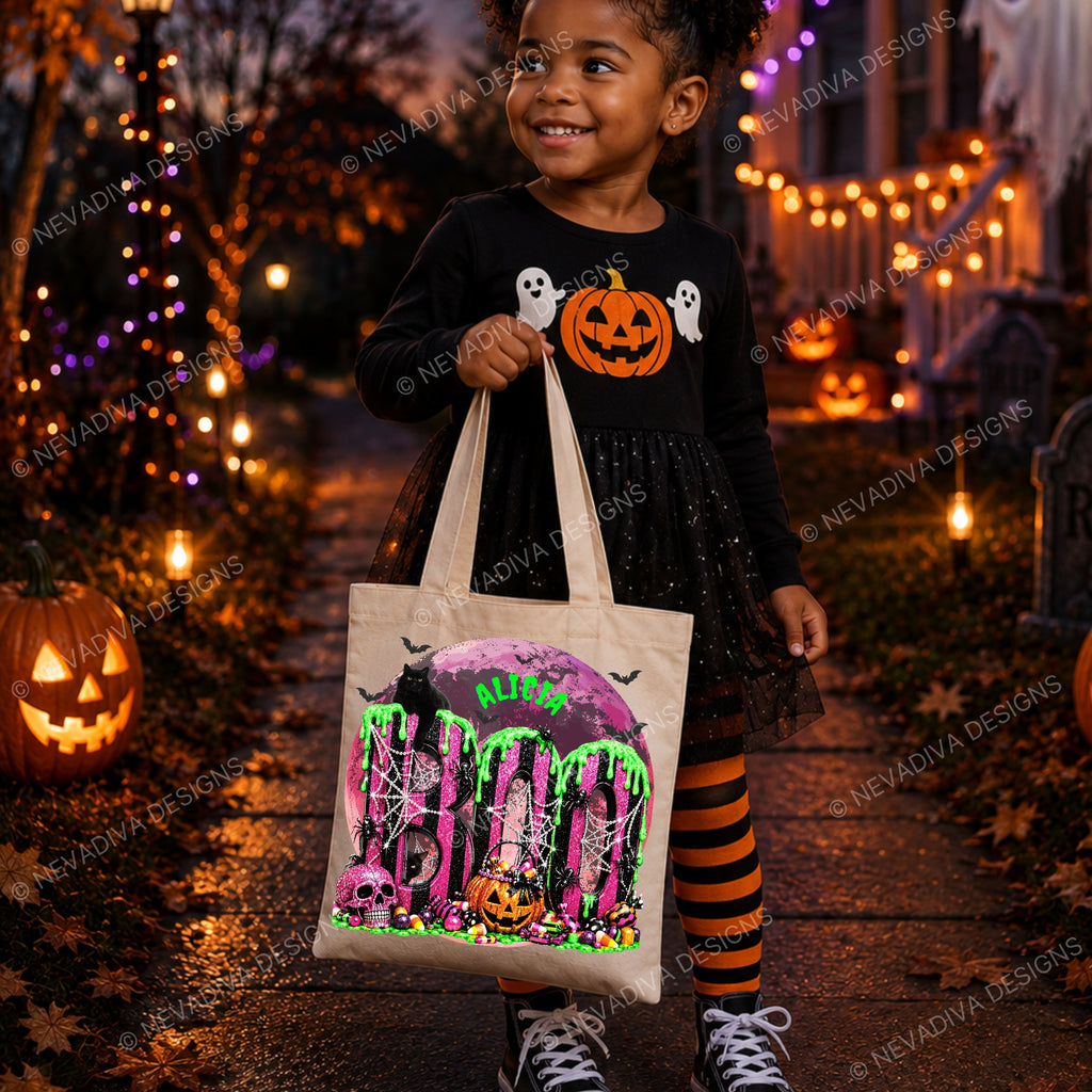 A young girl in a black Halloween dress and orange-and-black striped tights holds a cream canvas tote bag printed with a spooky 'BOO' design, standing on a decorated Halloween porch with pumpkins and string lights.