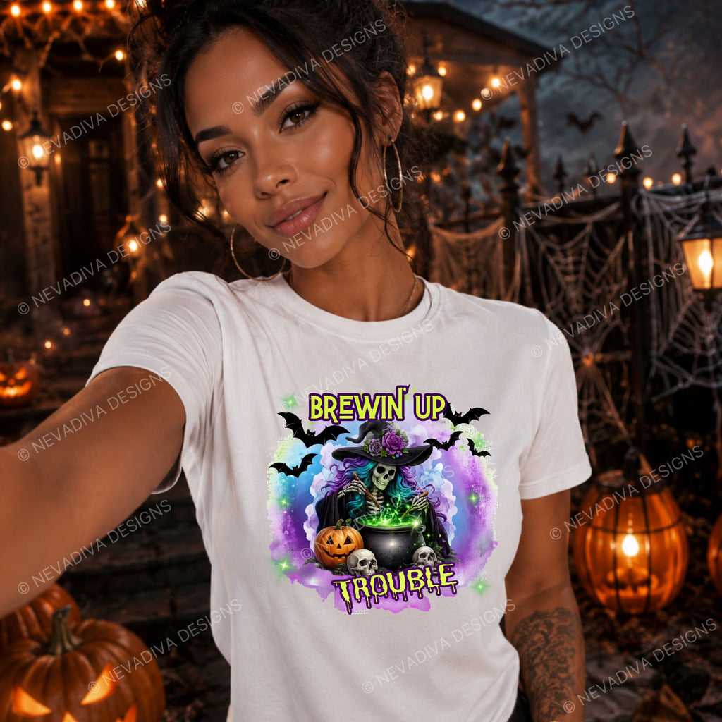 A woman wearing a white graphic t-shirt with a Halloween design that reads 'BREWIN' UP TROUBLE' around a witch, cauldron, and pumpkins. The background is a spooky Halloween scene with string lights, cobwebs, and pumpkins.