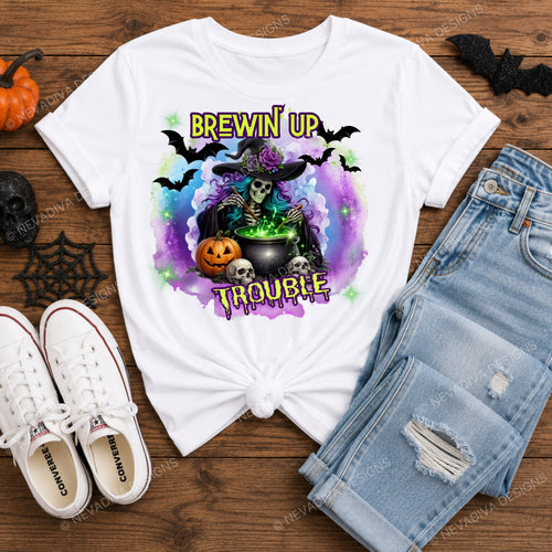 A white graphic T-shirt with a Halloween design featuring a skeleton witch, cauldron, pumpkins, bats, and the text 'BREWIN' UP TROUBLE', styled in a flat lay on a wooden surface with jeans, sneakers, and small Halloween props.