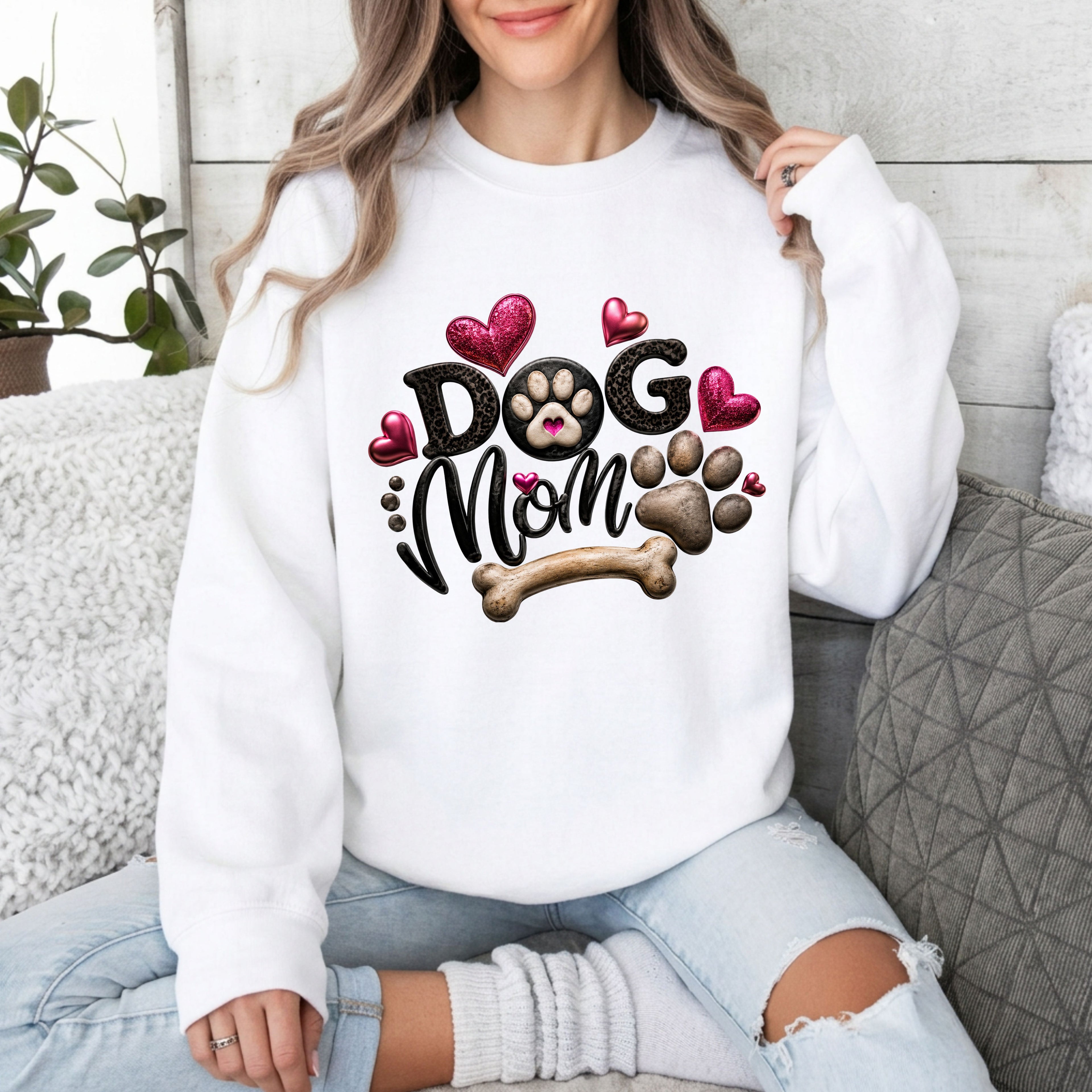 A woman wearing a white crewneck sweatshirt with a 'Dog Mom' graphic featuring hearts, a paw print, and a bone, sitting on a couch.