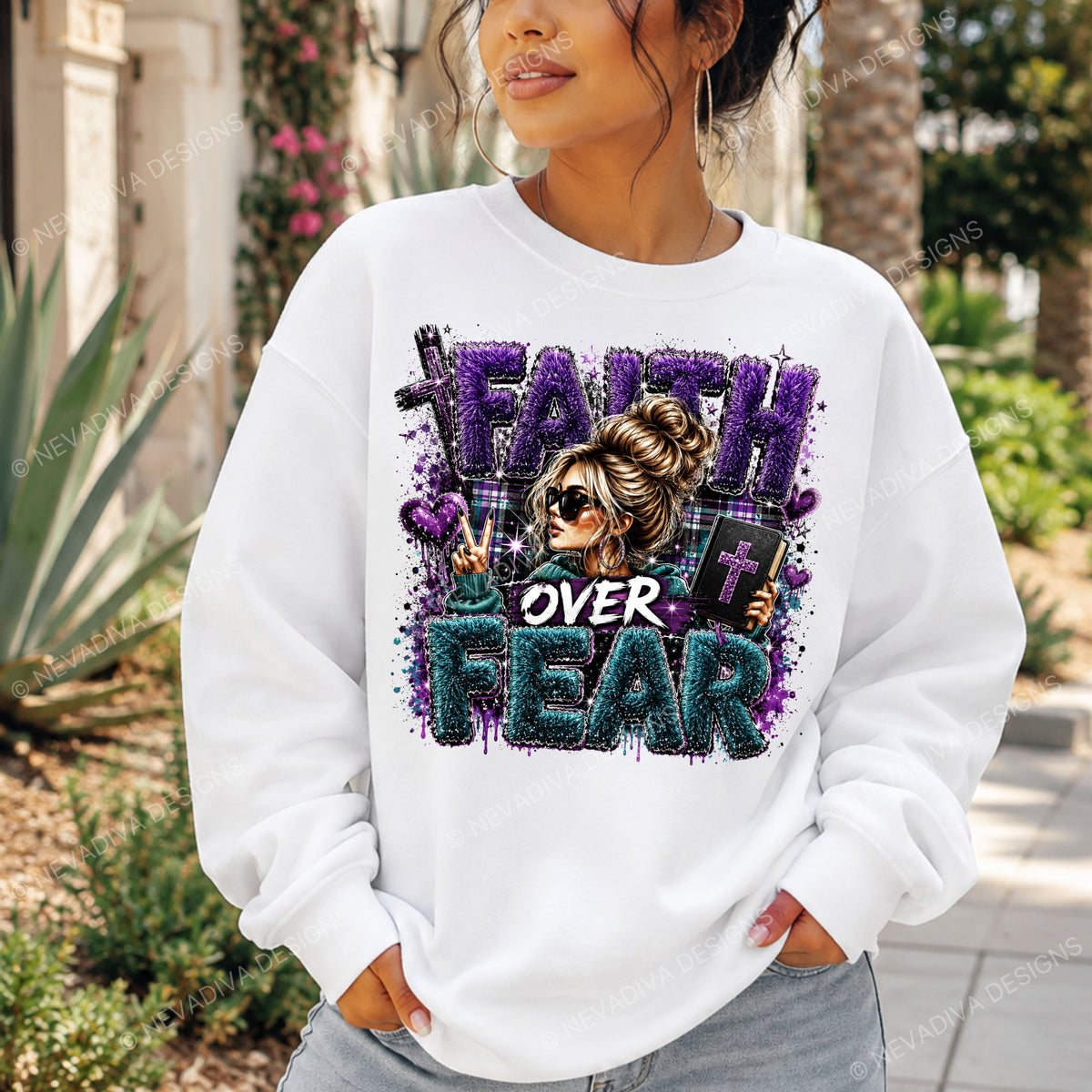 A woman wearing a white crewneck sweatshirt with a colorful graphic that reads 'FAITH OVER FEAR' in purple and teal glitter-style lettering, featuring an illustration of a woman with sunglasses. The photo is taken outdoors in a garden setting.