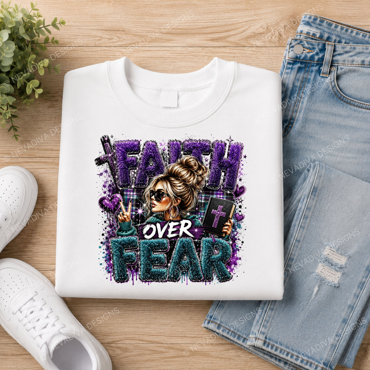 A folded white graphic T-shirt displayed on a wooden surface, styled with light blue jeans, white sneakers, and a small green plant. The shirt features a colorful graphic with the text 'FAITH OVER FEAR' and an illustrated woman holding a cross.
