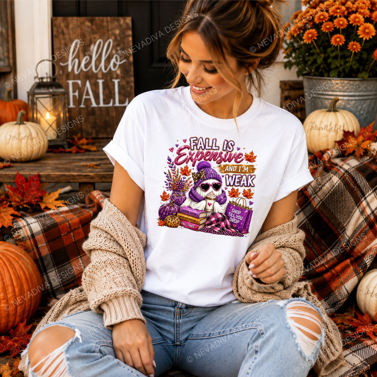 A woman wearing a white graphic t-shirt with a purple ghost and autumn-themed text, styled with a beige knit cardigan and ripped blue jeans in a cozy fall-themed setting.