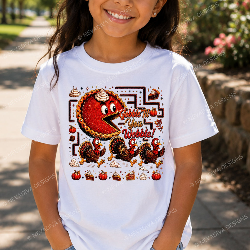 A child wearing a white graphic t-shirt featuring a 'Gobble Up Your Webbs!' Thanksgiving-themed design with a red pie character, turkeys, pumpkins, and pies, photographed outdoors on a sunny day.