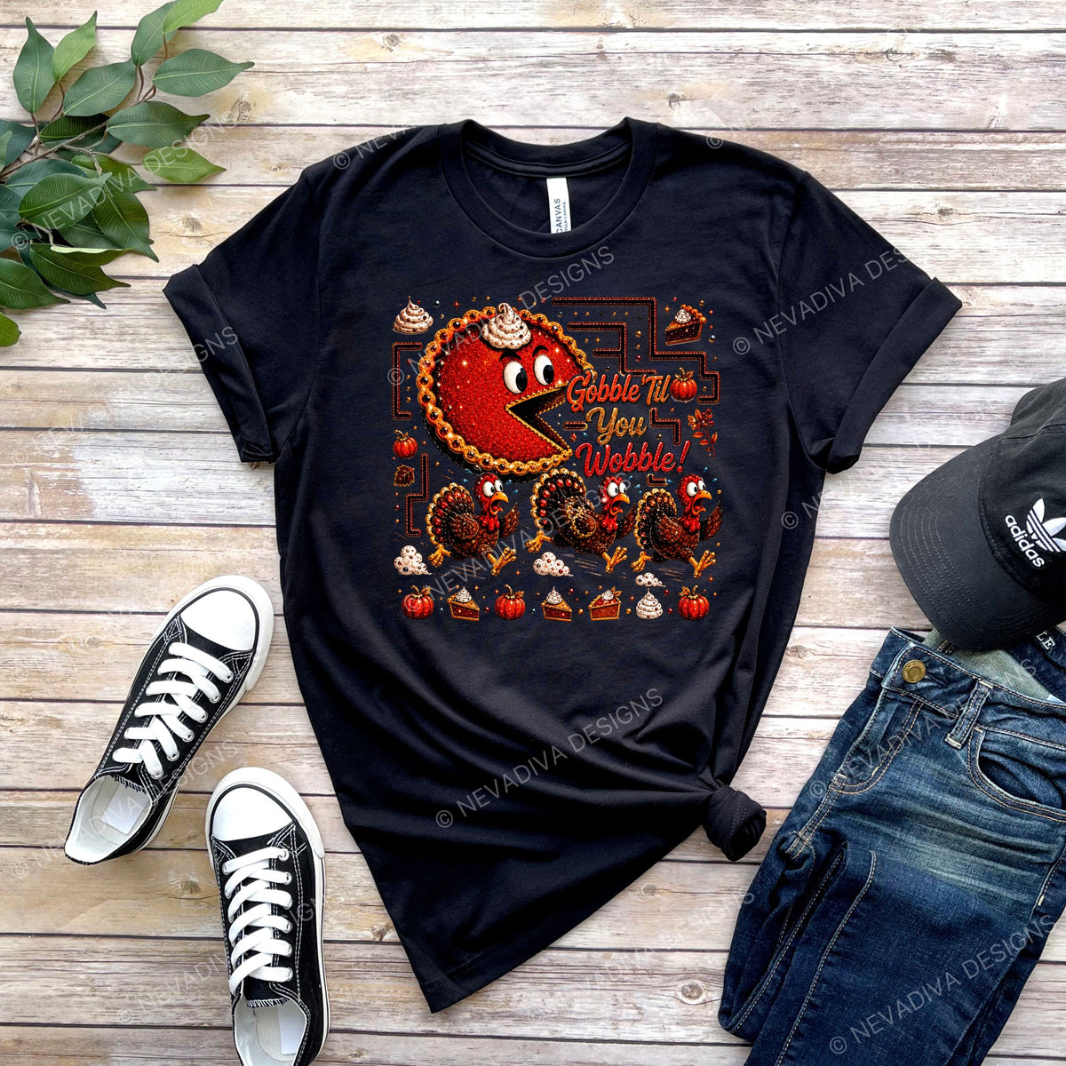 A black graphic t-shirt featuring a retro 'Gobble Up You Wobble!' Thanksgiving design with a red and orange Pac-Man style turkey and cupcakes, styled in a flat lay on a wooden background with black sneakers, blue jeans, and a black cap.