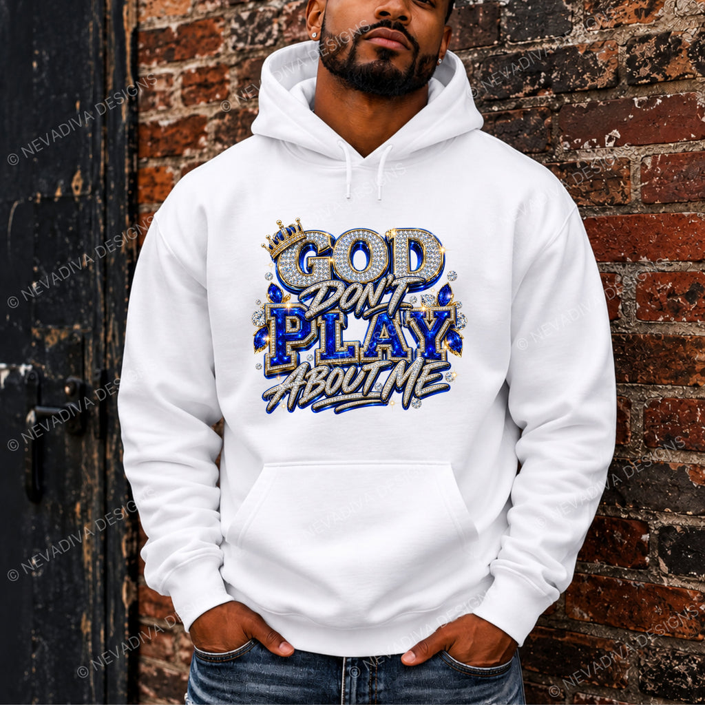 A man wearing a white pullover hoodie with a bold graphic that reads 'GOD DON'T PLAY ABOUT ME' in blue, gold, and white lettering, posed against a rustic brick wall.