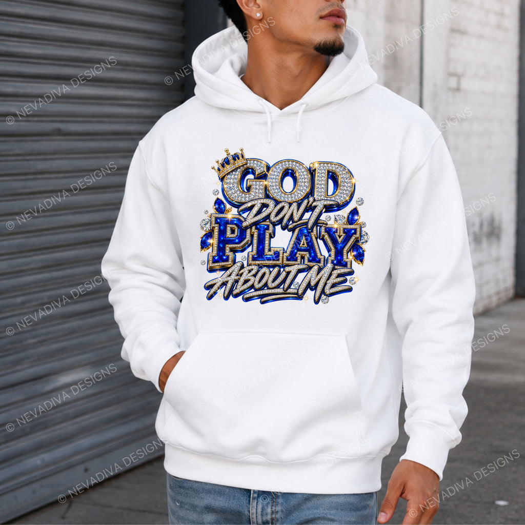 A man wearing a white pullover hoodie with a graphic that reads 'GOD DON'T PLAY ABOUT ME' in blue, gold, and white lettering, standing in front of a gray metal shutter and white brick wall.