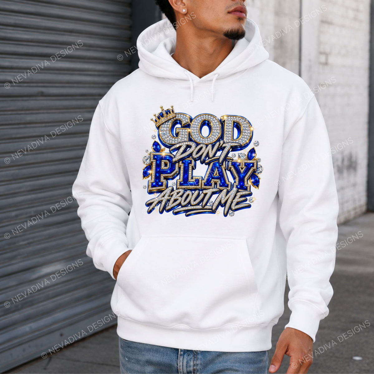 A man wearing a white pullover hoodie with a graphic that reads 'GOD DON'T PLAY ABOUT ME' in blue, gold, and white lettering, standing in front of a gray metal shutter and white brick wall.