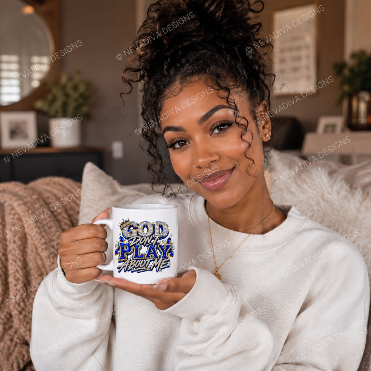 A woman in a white sweatshirt holds a white ceramic mug with a graphic that reads 'GOD IS NOT PLAYING WITH ME' in blue and yellow. The photo is taken in a cozy, modern living room setting with a white faux fur throw pillow behind her.