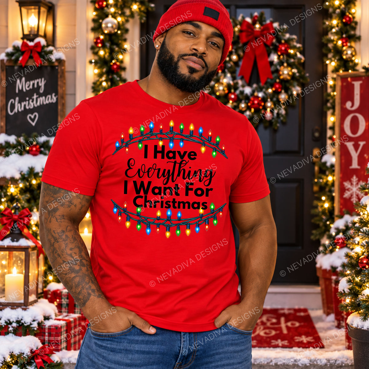 A man wearing a bright red graphic t-shirt and a red beanie stands in front of a decorated Christmas doorway. The t-shirt features a black graphic with the text 'I Have Everything I Want For Christmas' framed by colorful Christmas lights.