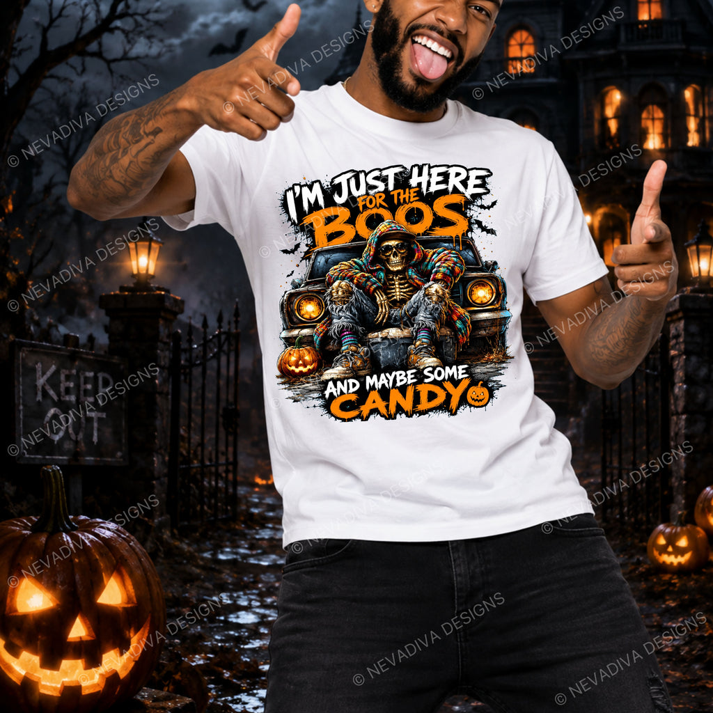 A man wearing a white graphic t-shirt with a Halloween design, posing in a spooky haunted house yard with pumpkins and lanterns. The t-shirt features a cartoon graphic of a skeleton in a candy corn hat and the text 'I'M JUST HERE FOR THE BOOS AND MAYBE SOME CANDY'.