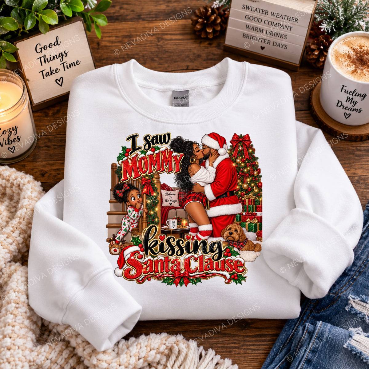 A white crewneck sweatshirt with a colorful Christmas graphic print, styled in a cozy flat-lay arrangement on a wooden table with a candle, card, mug, and denim jeans.