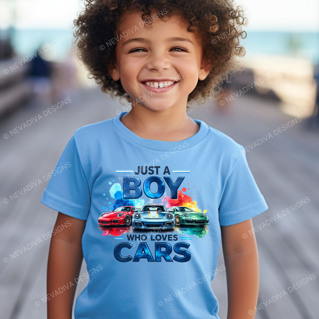 little boy wearing a blue short-sleeve t-shirt with a colorful graphic print on the front. The graphic features the text 'JUST A BOY WHO LOVES CARS' in stylized blue lettering above three sports cars in red, white, and green, set against a clean white background.