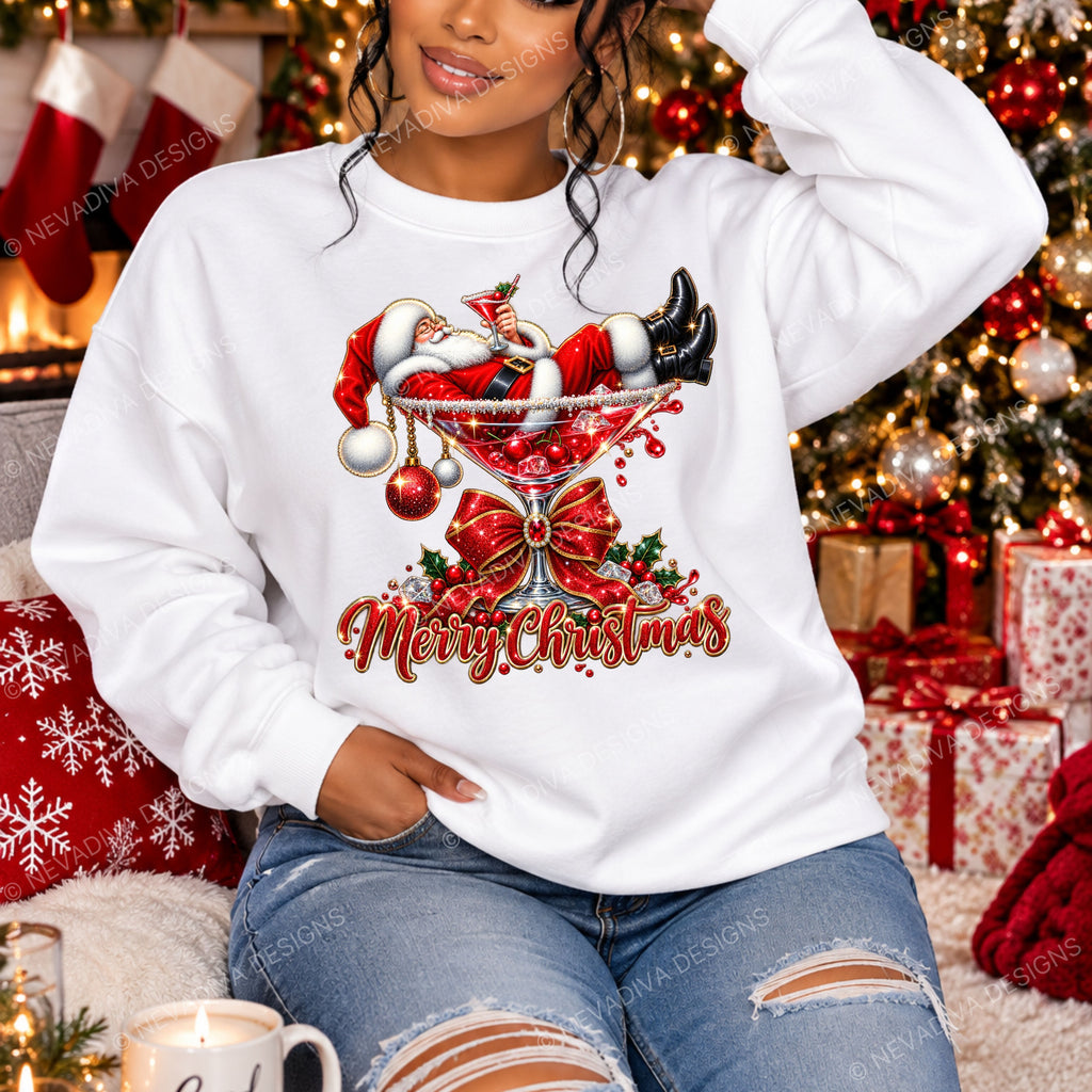 A woman wearing a white crewneck sweatshirt with a festive Christmas graphic of Santa Claus lounging in a martini glass and the words 'Merry Christmas' in red script. She is sitting in a cozy, decorated living room with a Christmas tree, stockings, and wrapped gifts in the background.