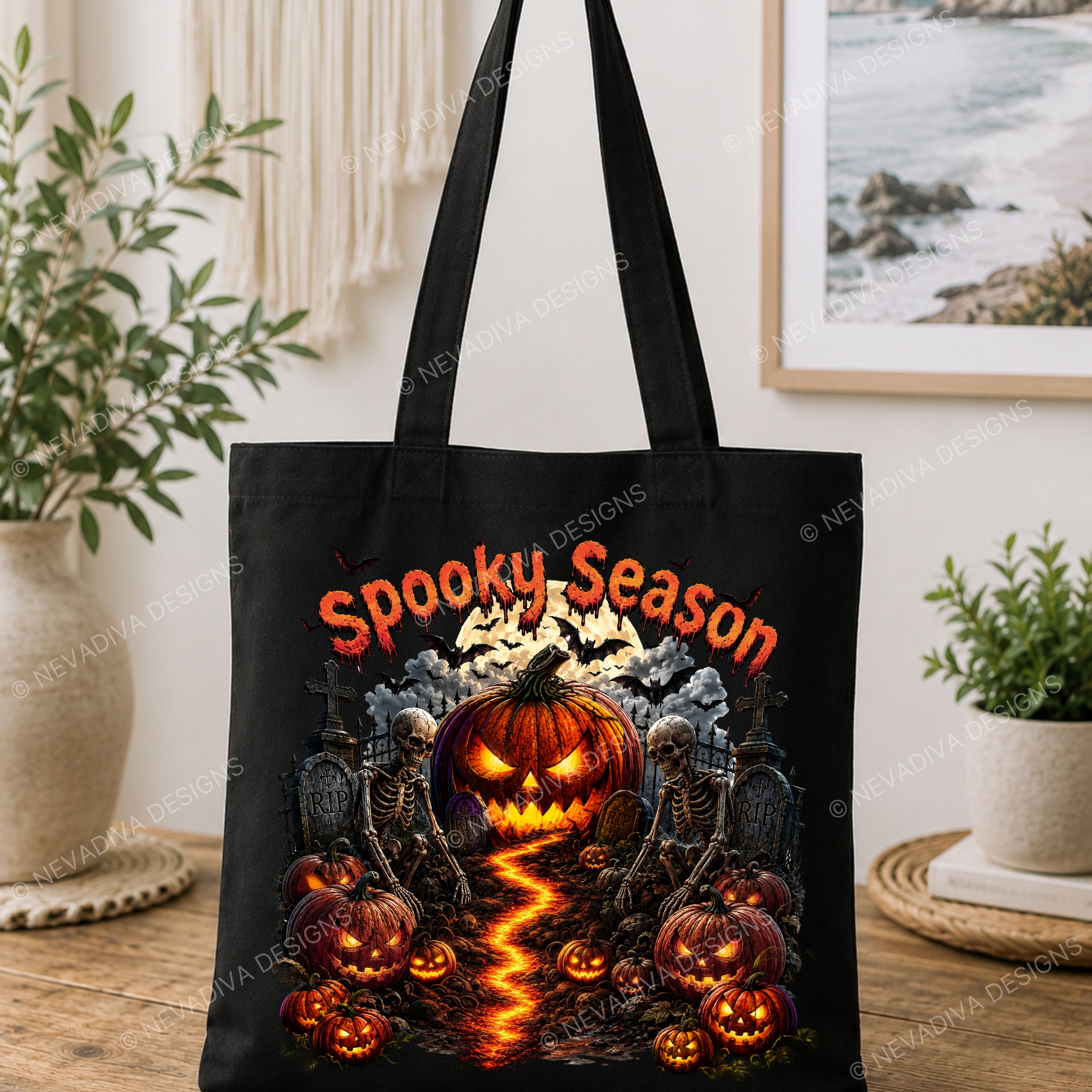 A black canvas tote bag with a detailed Halloween graphic print featuring a glowing jack-o'-lantern, skeletons, bats, pumpkins, and the text 'Spooky Season' in orange. The bag is displayed on a wooden table in a styled home interior setting with plants and a framed picture in the background.