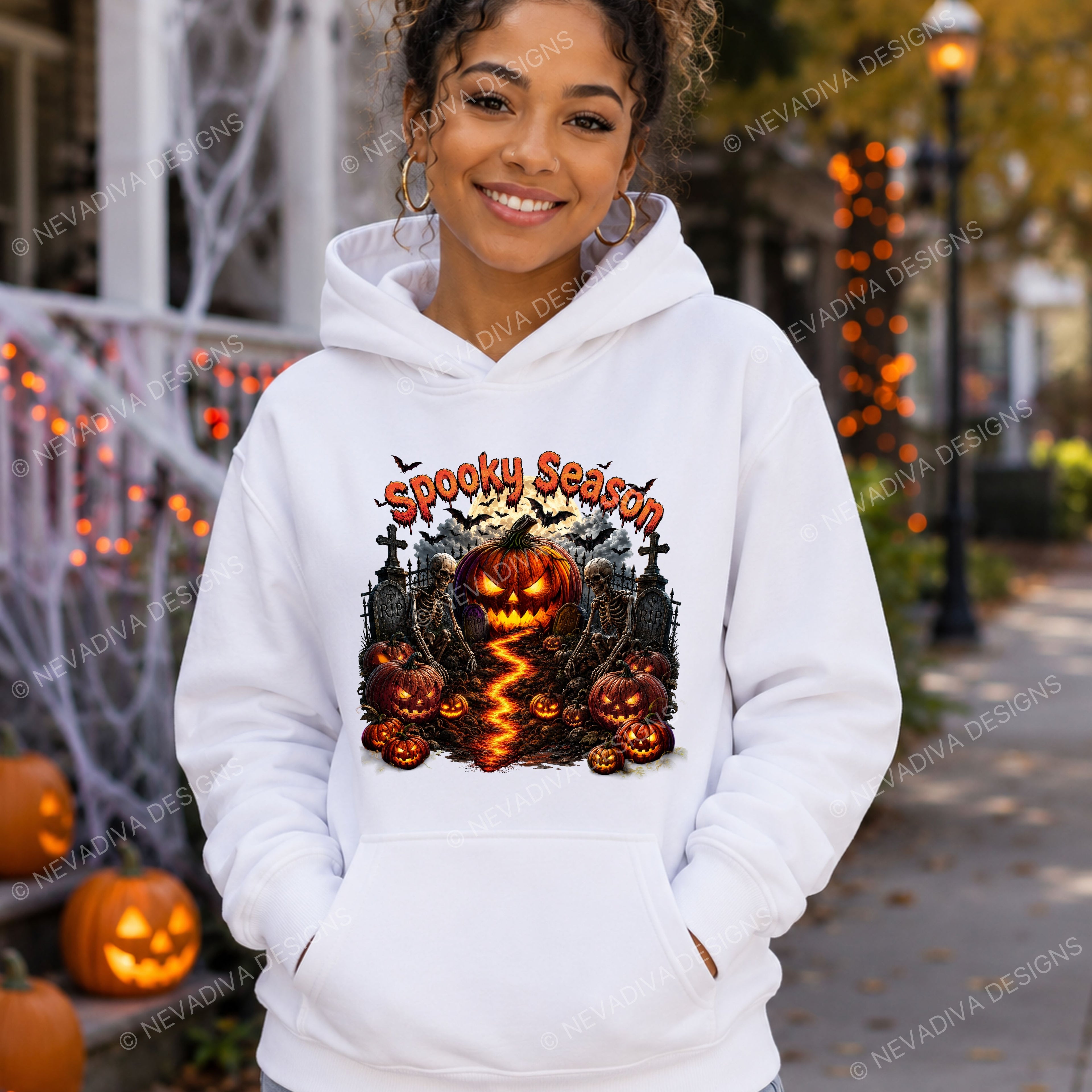 A woman wearing a white hoodie with a 'Spooky Season' Halloween graphic featuring jack-o'-lanterns, a haunted house, and lightning, standing outdoors in front of a decorated street scene.
