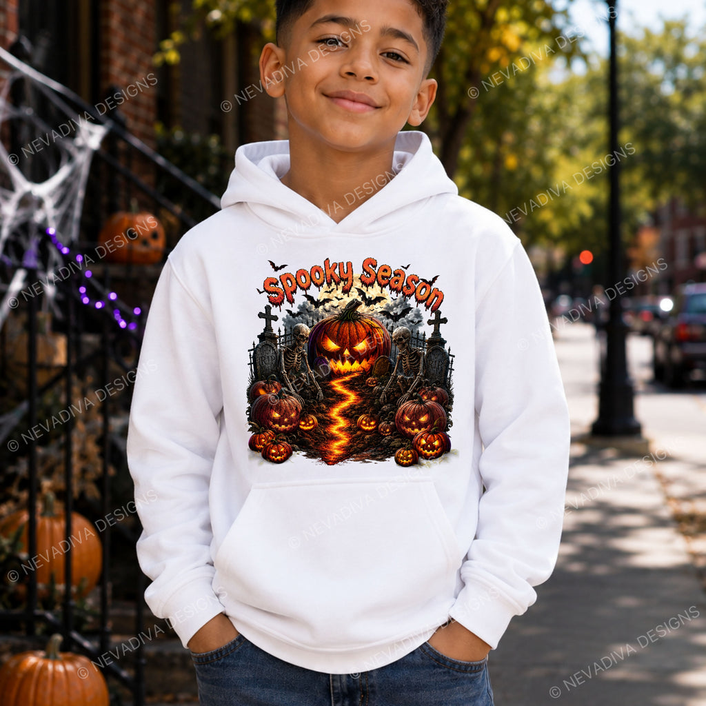 A young boy wearing a white hoodie with a 'Spooky Season' Halloween graphic featuring jack-o'-lanterns and a haunted house, standing on a city street with pumpkins and cobwebs in the background.