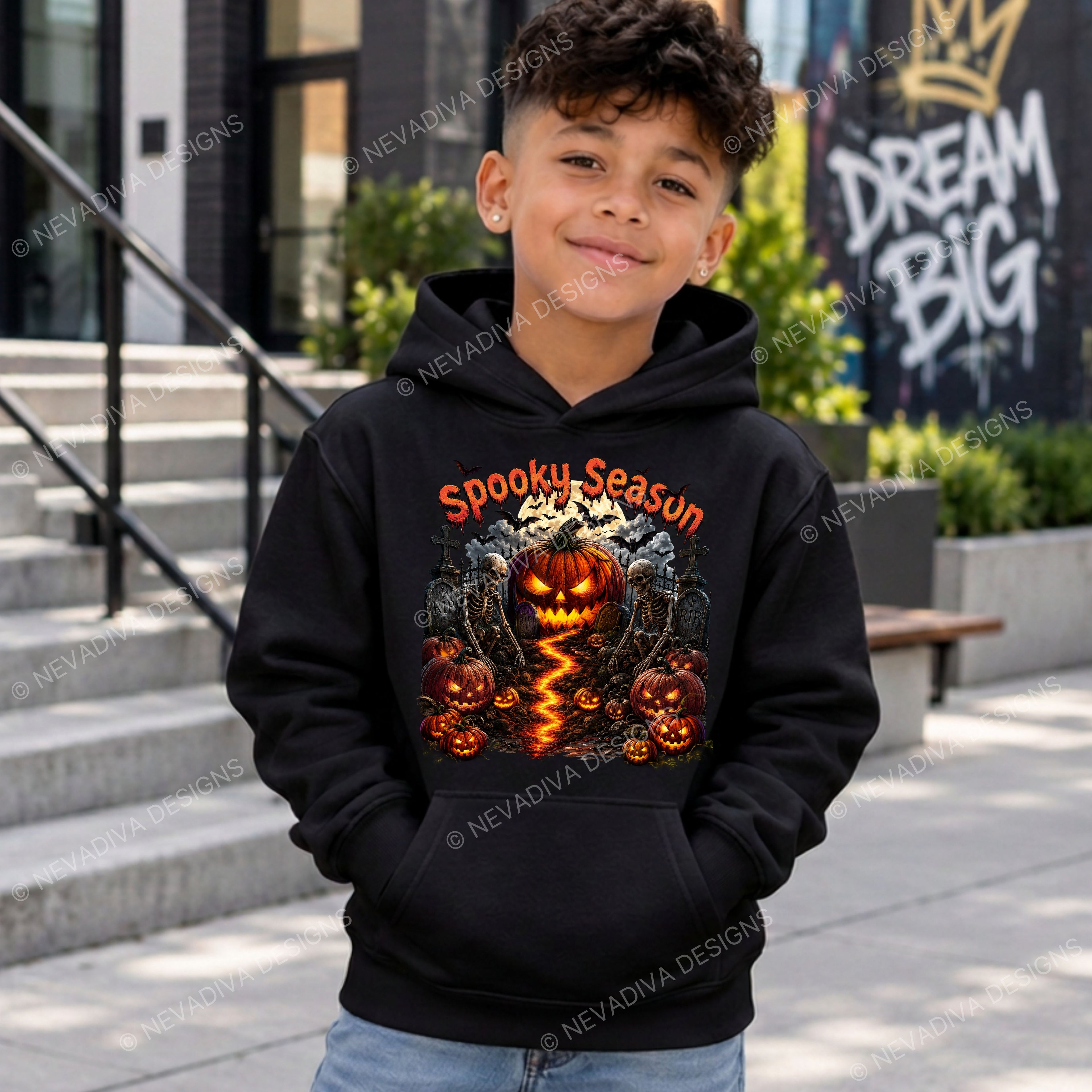 A young boy wearing a black hoodie with a 'Spooky Season' jack-o'-lantern graphic, standing outdoors in front of concrete steps and a building wall.