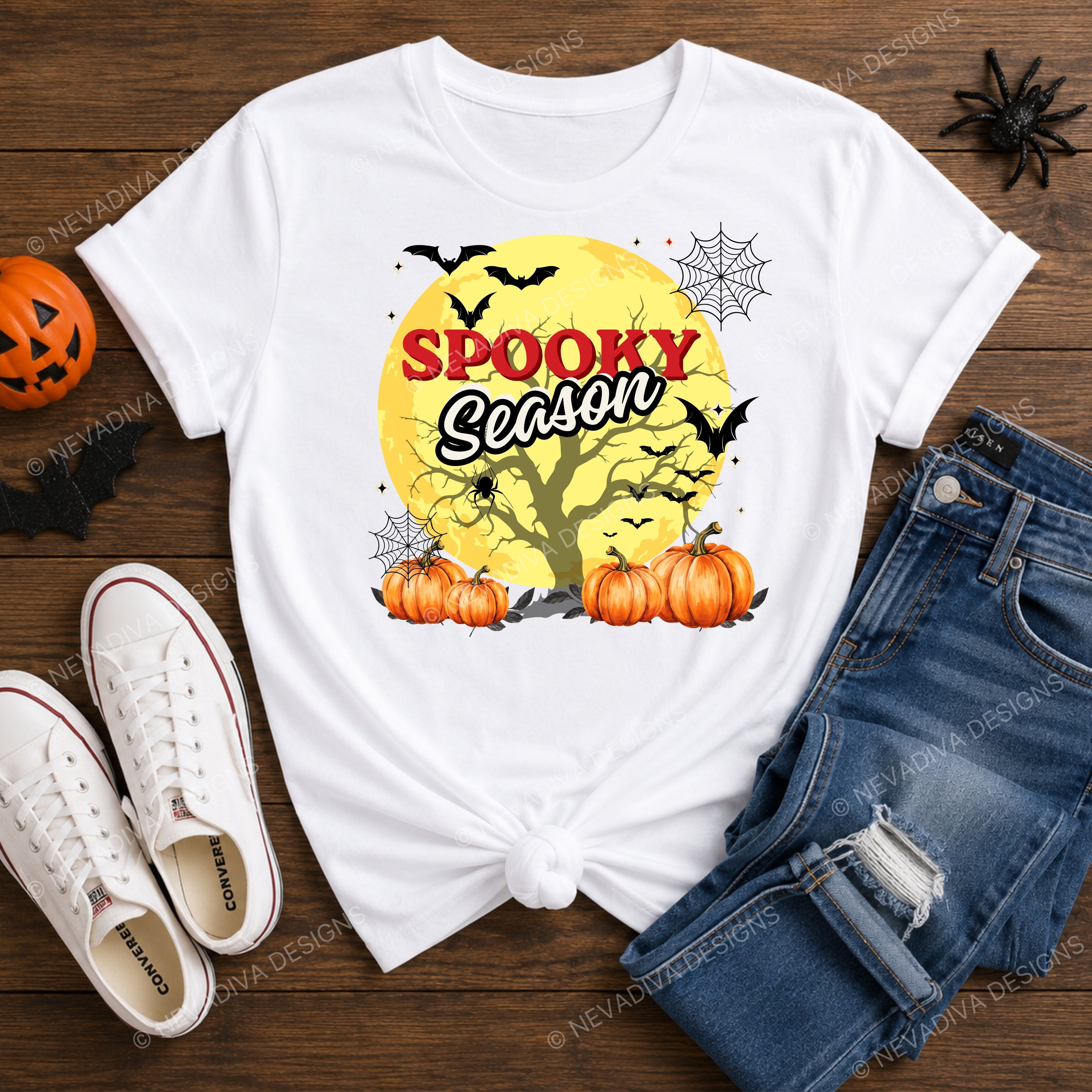 A white graphic t-shirt with a 'Spooky Season' Halloween design, styled in a flat lay on a wooden surface with jeans, white sneakers, a jack-o'-lantern, and a spider decoration.
