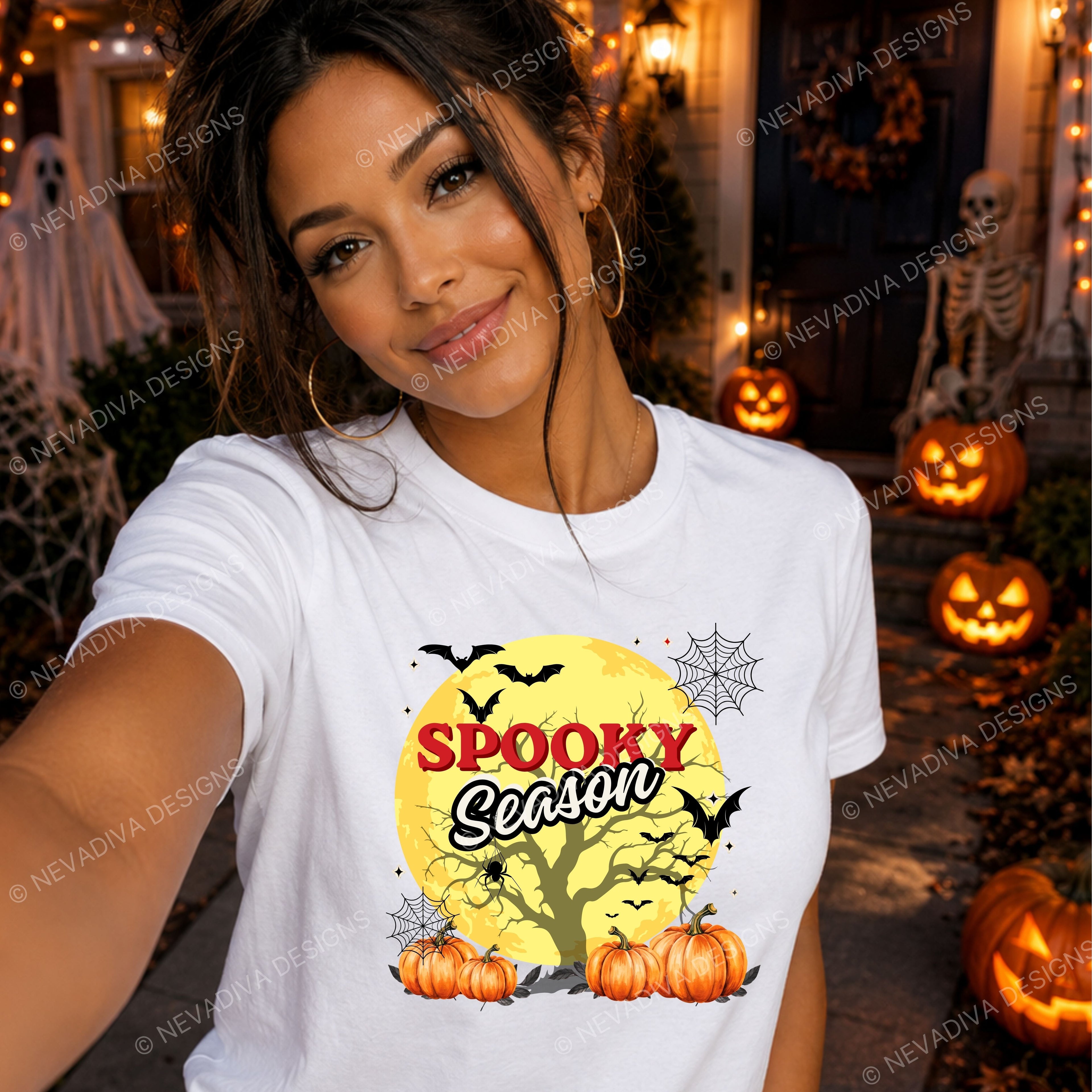 A woman takes a selfie wearing a white graphic t-shirt with a 'Spooky Season' Halloween design featuring pumpkins, a tree, bats, and a yellow moon. The background shows a decorated Halloween porch with jack-o'-lanterns, a skeleton, and string lights.