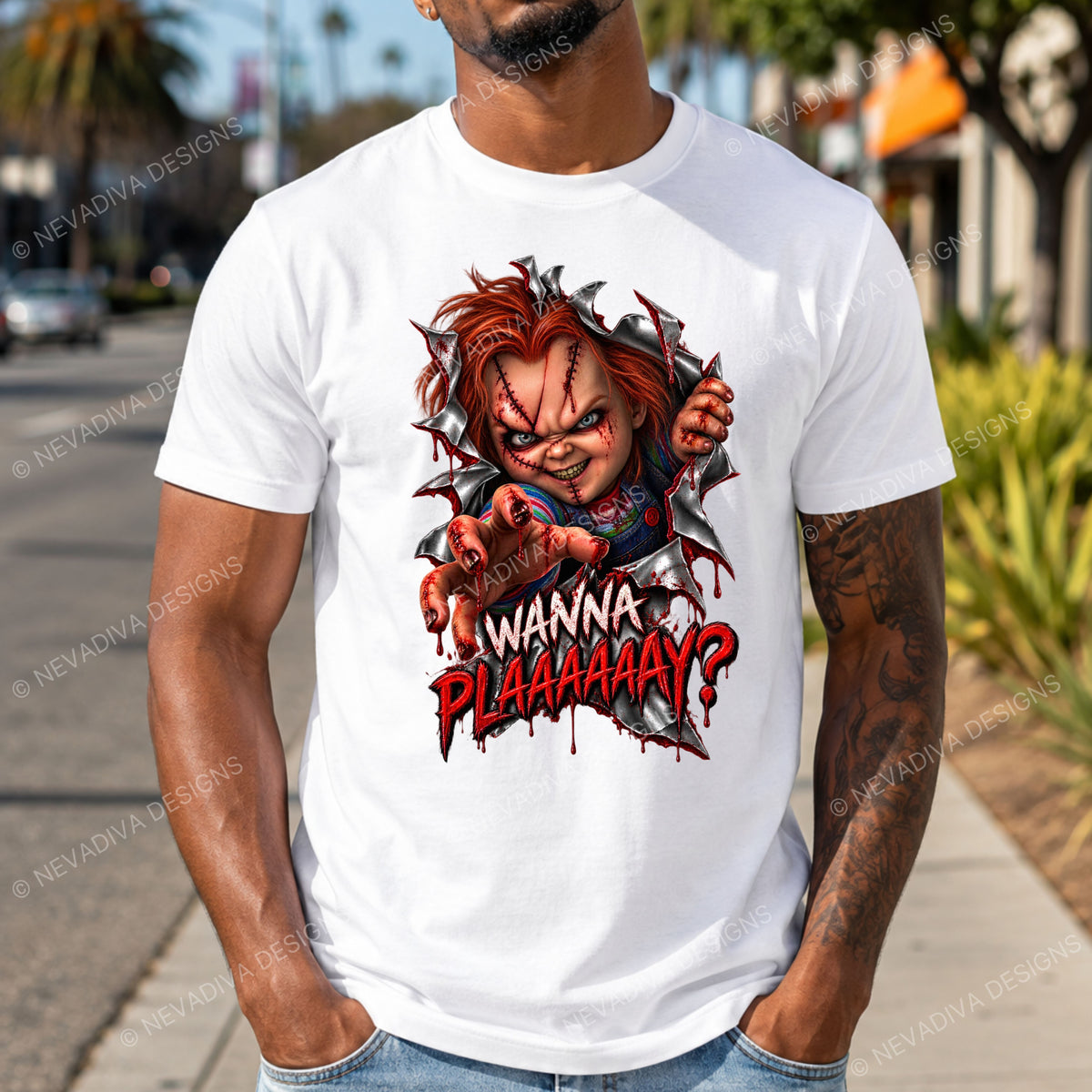 A man wearing a white graphic t-shirt featuring a stylized Chucky character design with the text 'Wanna Plaaaay?'. The background is a blurred outdoor street scene.