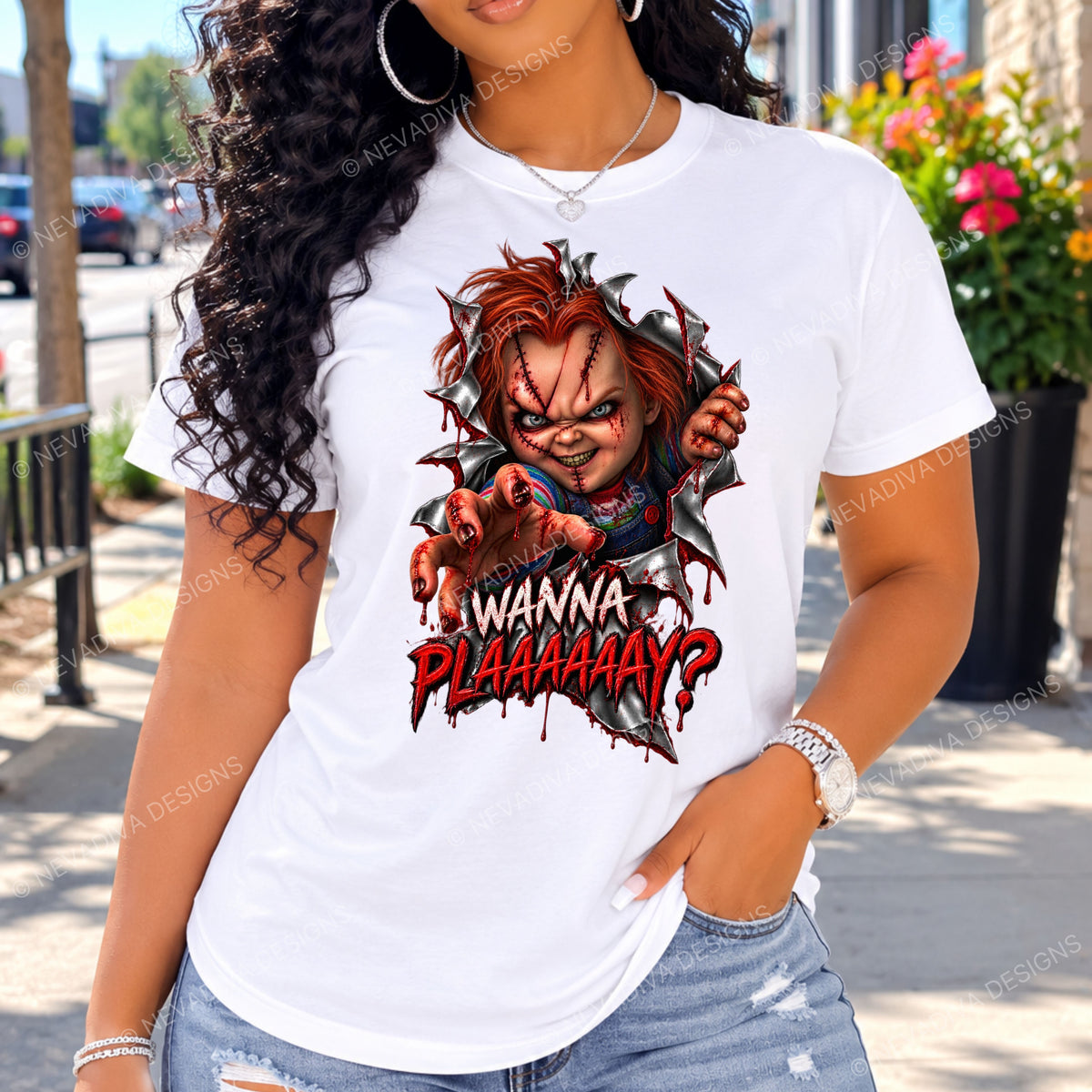 A woman wearing a white graphic t-shirt featuring a stylized Chucky character tearing through the fabric with the text 'I WANNA PLAAAAAAAY?' in red and black. The background shows an outdoor street scene with blurred elements.