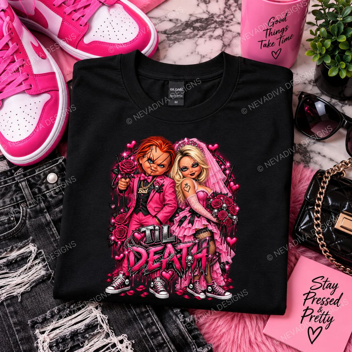 A folded black graphic T-shirt is displayed in a styled flat-lay with pink sneakers, ripped denim, sunglasses, a pink cup, a small plant, and a black quilted handbag around it. The shirt features a large pink and multicolor graphic of two cartoon characters with roses and the text 'Till Death'.