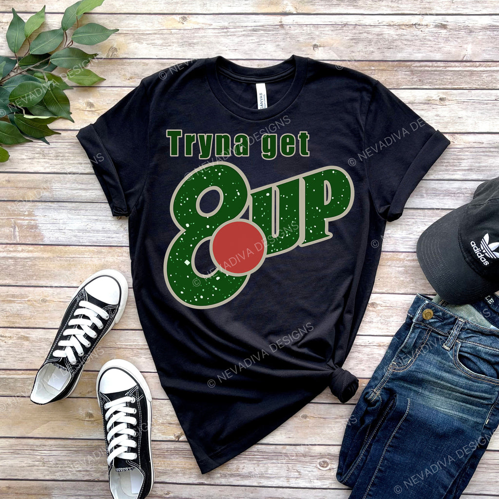 Black t-shirt with 'Tryna get 8 up' design on a wooden surface with shoes and jeans.