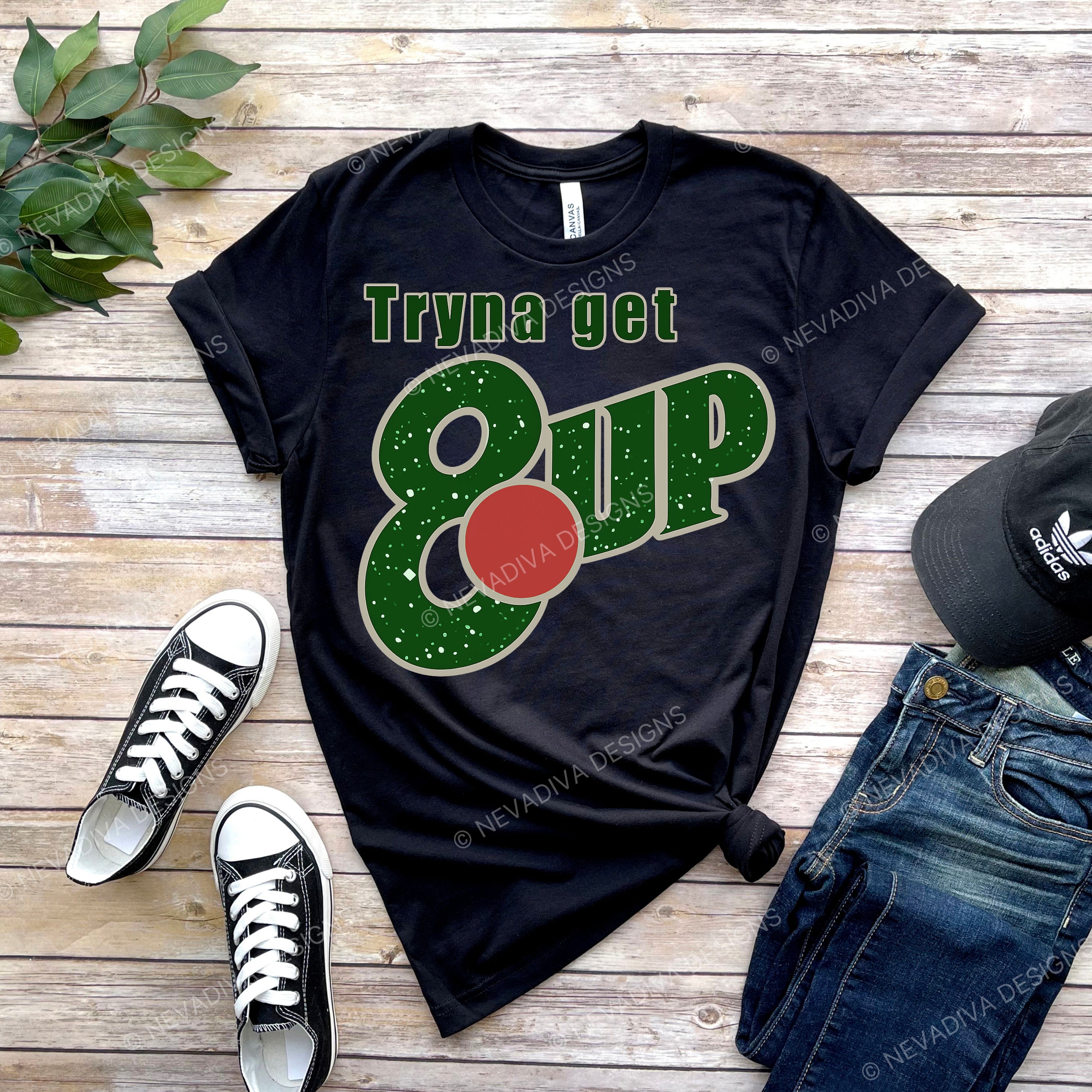Black t-shirt with 'Tryna get 8 up' design on a wooden surface with shoes and jeans.