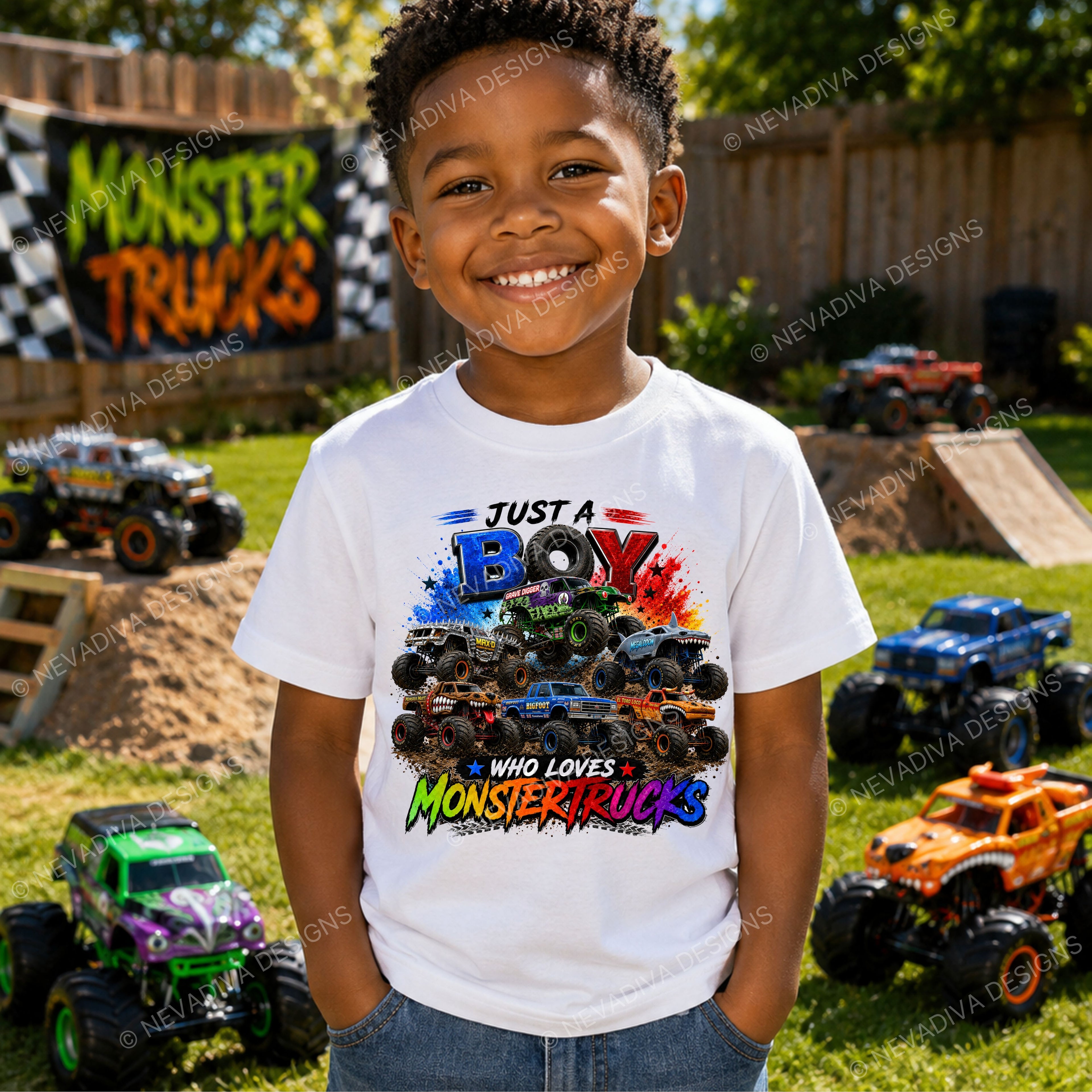 A smiling young boy stands outdoors in a backyard play area wearing a white graphic t-shirt with a colorful monster truck print that reads 'JUST A BOY WHO LOVES MONSTER TRUCKS'. Several monster trucks are parked on the grass and dirt ramps in the background, with a wooden fence and trees behind him.