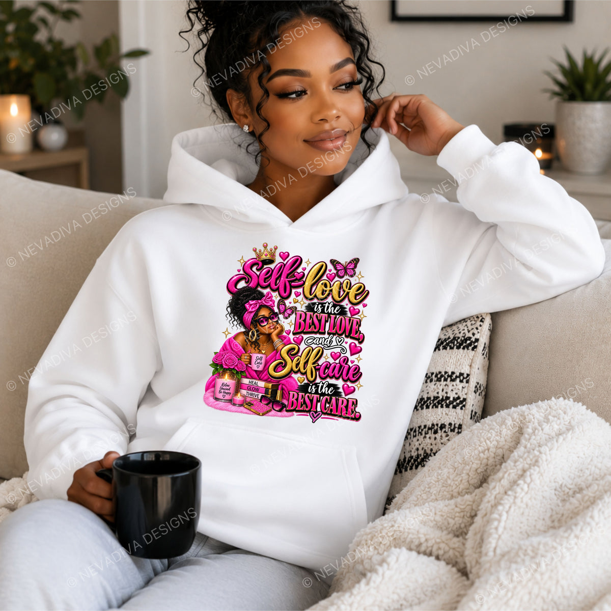 A woman sits on a beige couch wearing a white hoodie with a colorful graphic print that reads 'Self-love is the best love, and Self-care is the best care' with a pink illustrated figure. She holds a black mug in a cozy living room setting.