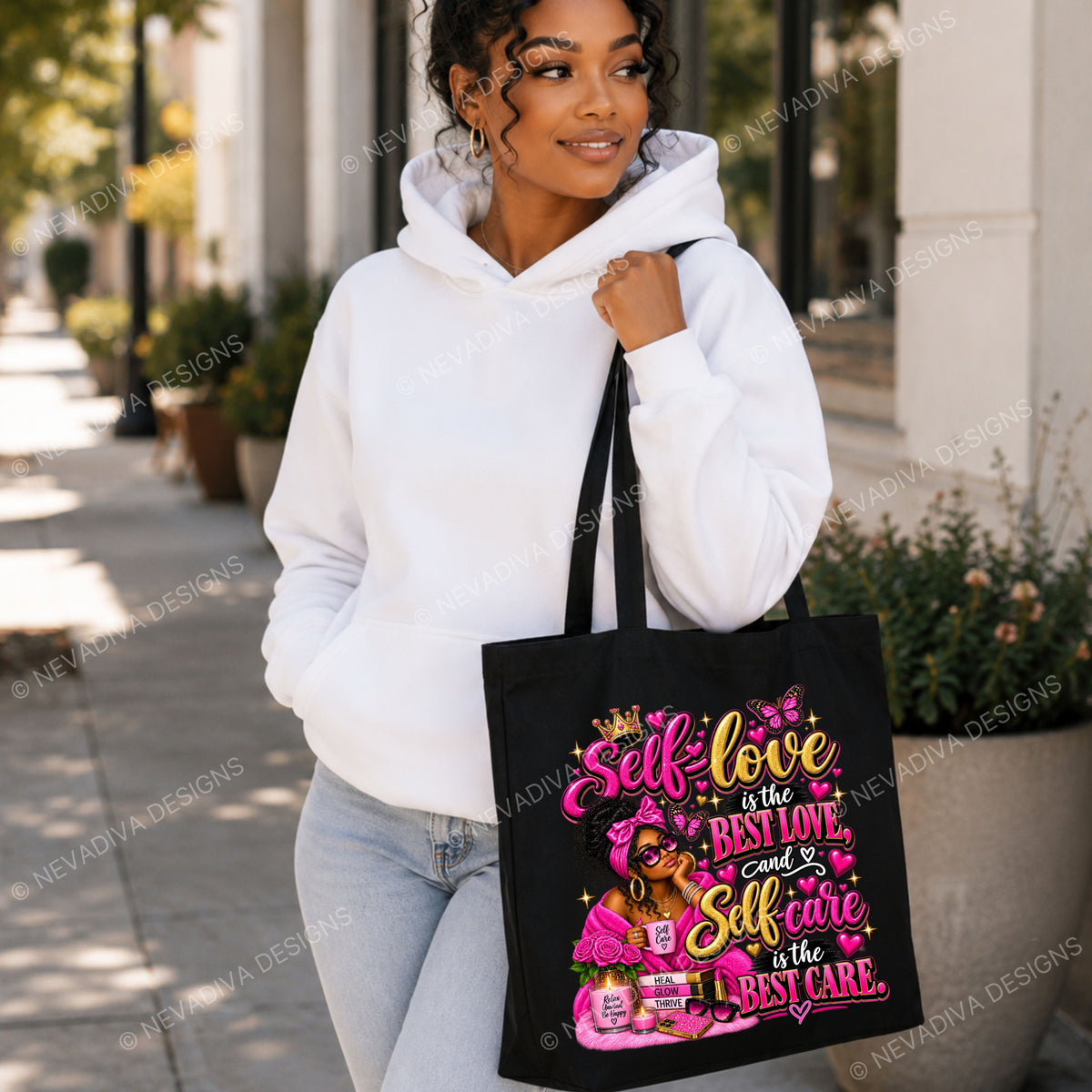 A woman wearing a white hoodie and light blue jeans stands on a city sidewalk holding a black tote bag with a colorful graphic that reads 'Self love is the best love, and self care is the best care' with pink and gold lettering and illustrations.